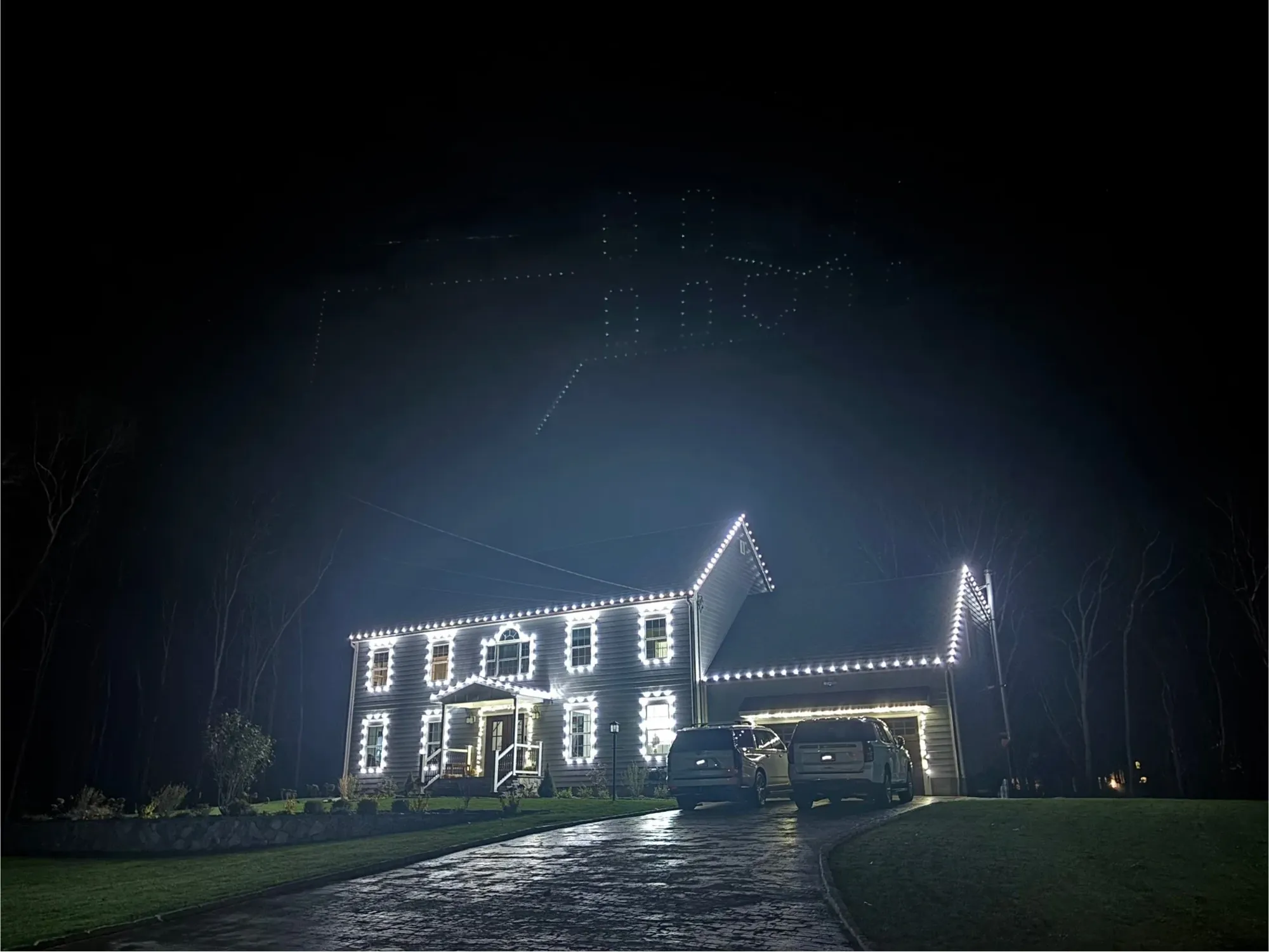 Two-story colonial with a lit roofline, window frames and garage on a long driveway